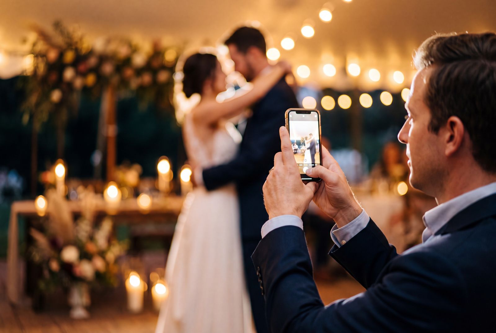 A wedding guest filming the couple's first dance on a phone, warm candlelight behind them